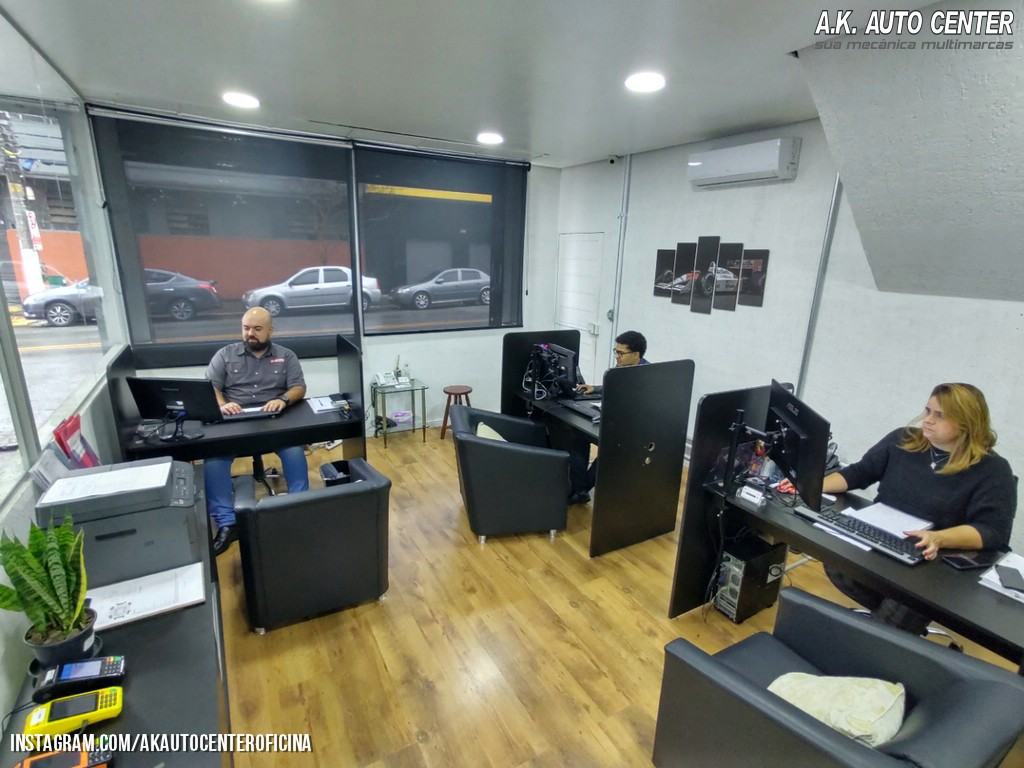 Recepção da AK Auto Center com piso de madeira, três atendentes em mesas com computadores e poltronas para quem espera.